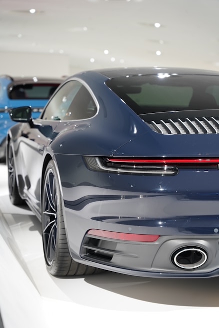 A sleek, dark blue sports car is prominently displayed, focusing on the rear end. The vehicle features a modern design with a distinctive rear light bar and dual exhaust. The setting appears to be indoors, possibly a showroom, with a soft, bright lighting ambiance.