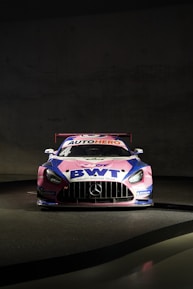 A race car is prominently displayed against a dark background. The vehicle is primarily pink, blue, and white, featuring branding from 'BWT' and 'AUTOHERO' on the hood and windshield. The car has a distinctive aerodynamic design typical of racing cars, with a prominent front grille and sleek headlights. Mercedes-Benz and Michelin logos are also visible on the car.