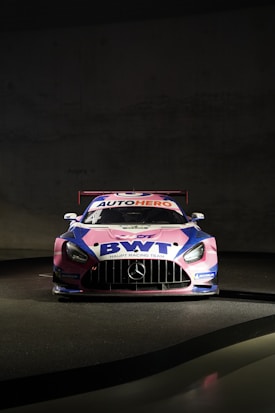 A race car is prominently displayed against a dark background. The vehicle is primarily pink, blue, and white, featuring branding from 'BWT' and 'AUTOHERO' on the hood and windshield. The car has a distinctive aerodynamic design typical of racing cars, with a prominent front grille and sleek headlights. Mercedes-Benz and Michelin logos are also visible on the car.