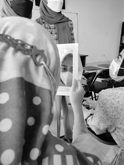 People wearing headscarves and masks are gathered in a room. One person is holding a mirror reflecting part of a face. Chairs and a whiteboard can be seen in the background.