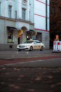 A white taxi with branding on its side is parked on a street in front of a building with large windows and decorative moldings. The street is lined with autumn leaves, and there is a small barricade with a warning light.