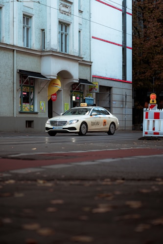 A white taxi with branding on its side is parked on a street in front of a building with large windows and decorative moldings. The street is lined with autumn leaves, and there is a small barricade with a warning light.