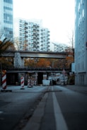 A city street is lined with construction barriers and signs, suggesting road work or an ongoing project. The street is bordered by tall residential buildings, and a pedestrian overpass extends above the road. The surrounding trees display autumn colors.