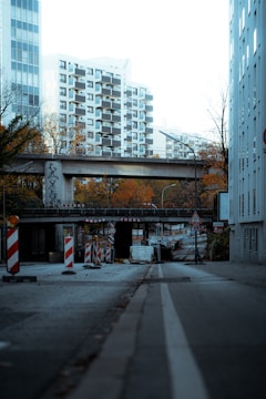 A city street is lined with construction barriers and signs, suggesting road work or an ongoing project. The street is bordered by tall residential buildings, and a pedestrian overpass extends above the road. The surrounding trees display autumn colors.