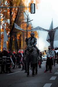 A group of people wearing dark, post-apocalyptic costumes are gathered on a street. The focal point is a figure in armor mounted on a dark horse, holding a lance with abstract geometric structures. Surrounding them are individuals with red face coverings, some carrying banners and other symbolic objects. The setting includes autumnal trees with orange foliage, adding contrast to the somber attire.