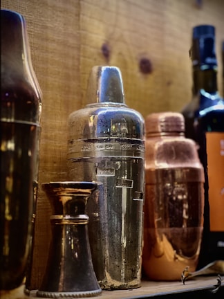 A collection of vintage cocktail shakers and bar tools arranged on a wooden shelf. The shakers are made of different materials, including metal and copper, each with a unique patina showcasing wear and age. A few bottles are visible in the background, adding to the rustic bar atmosphere.
