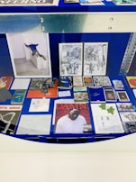 A display case with a variety of artworks and printed materials, including books, photographs, and album covers. A prominent album cover features a person wearing a white shirt against a red brick wall. There are black and white illustrations depicting urban scenes and abstract geometric drawings. The background is a deep blue, and there are metal edges visible.