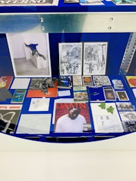 A display case with a variety of artworks and printed materials, including books, photographs, and album covers. A prominent album cover features a person wearing a white shirt against a red brick wall. There are black and white illustrations depicting urban scenes and abstract geometric drawings. The background is a deep blue, and there are metal edges visible.