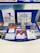 A display case with a variety of artworks and printed materials, including books, photographs, and album covers. A prominent album cover features a person wearing a white shirt against a red brick wall. There are black and white illustrations depicting urban scenes and abstract geometric drawings. The background is a deep blue, and there are metal edges visible.