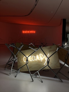 The image features a dimly lit room with a modern installation consisting of a wooden board displaying the word 'Occupy' in bright neon lights. In the background, a red neon sign reads 'Too late for tomorrow'. The installation is surrounded by metal stands, contributing to a minimalistic and industrial aesthetic.