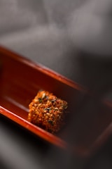 A small, cubed piece of food covered in spices and herbs is placed in the center of a wooden dish. The focus is on the vibrant texture and colors of the food, with a blurred background adding depth.