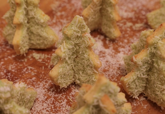 Festive tree-shaped cookies covered in shredded coconut are arranged on a wooden surface, creating a holiday theme. The cookies are light green with a hint of brown from baking.