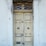 A weathered wooden double door with peeling paint, adorned with decorative metalwork above. Signs of decay are evident with chipped paint and worn surfaces surrounding the door frame.