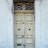 A weathered wooden double door with peeling paint, adorned with decorative metalwork above. Signs of decay are evident with chipped paint and worn surfaces surrounding the door frame.