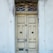 A weathered wooden double door with peeling paint, adorned with decorative metalwork above. Signs of decay are evident with chipped paint and worn surfaces surrounding the door frame.