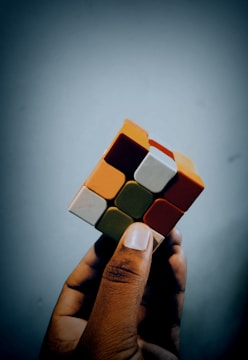 A person holds a partially solved Rubik's Cube against a plain background. The cube features various colored squares, including orange, white, red, and green.