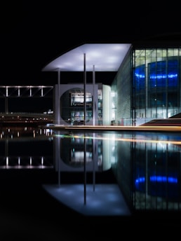 A modern architectural structure is beautifully lit at night, featuring large glass facades and geometric patterns, with lights reflecting on a body of still water in the foreground. The building has a distinctive circular design element and long vertical columns supporting a flat roof.