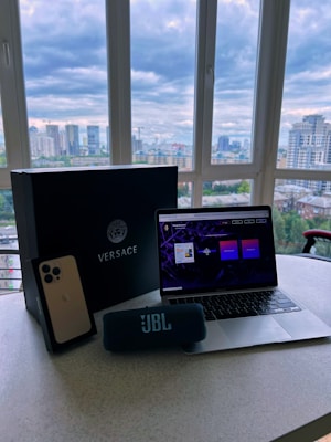 A modern setup featuring a laptop with an open screen, placed on a table. Beside the laptop, there is a Versace box, a smartphone positioned upright, and a JBL portable speaker. The table is situated in front of a large window with a view of a city skyline under a cloudy sky.