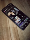 A smartphone is lying on a wooden surface, displaying a music streaming app that shows playlists and songs on the screen.
