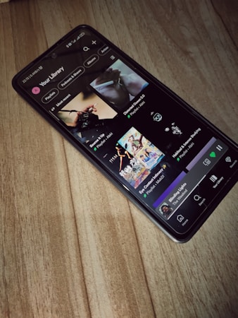 A smartphone is lying on a wooden surface, displaying a music streaming app that shows playlists and songs on the screen.