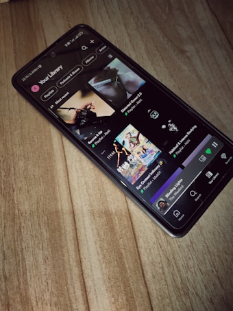 A smartphone is lying on a wooden surface, displaying a music streaming app that shows playlists and songs on the screen.