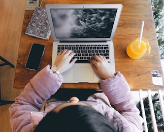 A person is typing on a laptop placed on a wooden table. Next to the laptop are a smartphone, a floral-patterned notebook, and a glass of orange juice with a straw. The person is wearing a striped pinkish-purple jacket and a wristwatch.