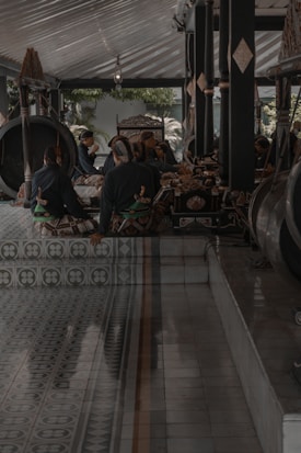 A group of individuals is seated in a traditional setting with musical instruments around them. The ornate decorations and patterned flooring contribute to the cultural atmosphere. The dim lighting and shadows create a solemn ambiance.