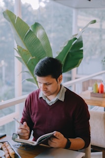 A person is sitting at a table in a bright, natural light setting, writing in a notebook with a pen. Large green leaves can be seen in the background, suggesting an indoor plant-filled area. The person is wearing a maroon sweater over a collared shirt and is focused on their writing.