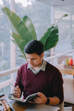 A person is sitting at a table in a bright, natural light setting, writing in a notebook with a pen. Large green leaves can be seen in the background, suggesting an indoor plant-filled area. The person is wearing a maroon sweater over a collared shirt and is focused on their writing.