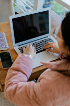 A friendly professional woman typing on a laptop with a bright pink background.