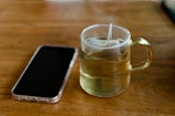 A clear glass cup filled with hot tea sits on a wooden table. The teabag string hangs over the edge of the cup. Next to it lies a smartphone with a translucent case, displaying its back side. The setting is simple and cozy, evoking a moment of pause or contemplation.