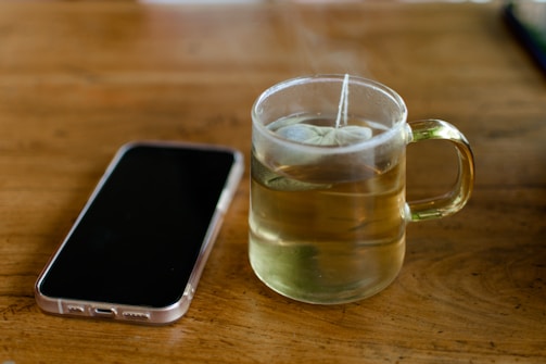 A clear glass cup filled with hot tea sits on a wooden table. The teabag string hangs over the edge of the cup. Next to it lies a smartphone with a translucent case, displaying its back side. The setting is simple and cozy, evoking a moment of pause or contemplation.