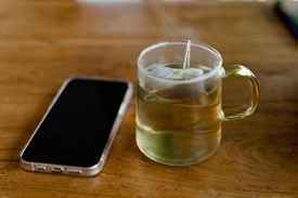 A clear glass cup filled with hot tea sits on a wooden table. The teabag string hangs over the edge of the cup. Next to it lies a smartphone with a translucent case, displaying its back side. The setting is simple and cozy, evoking a moment of pause or contemplation.