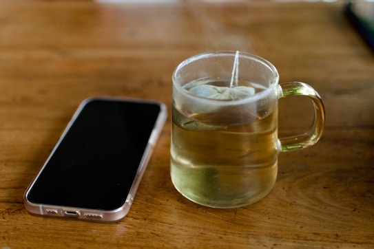 A clear glass cup filled with hot tea sits on a wooden table. The teabag string hangs over the edge of the cup. Next to it lies a smartphone with a translucent case, displaying its back side. The setting is simple and cozy, evoking a moment of pause or contemplation.