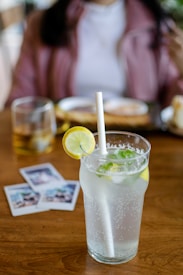 A refreshing glass of lemon-lime soda with ice, garnished with a slice of lemon and mint leaves, placed on a wooden table. Behind the drink, there are three instant photos and a glass with a beverage, along with a blurred view of a person in a casual setting.