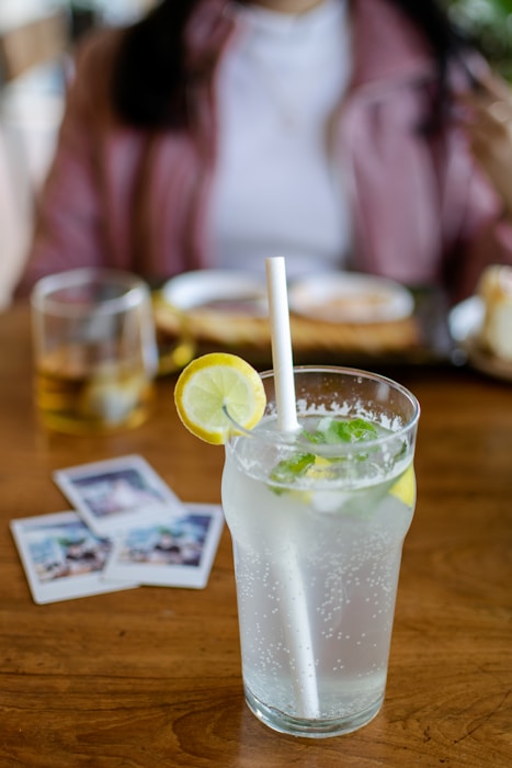 A refreshing glass of lemon-lime soda with ice, garnished with a slice of lemon and mint leaves, placed on a wooden table. Behind the drink, there are three instant photos and a glass with a beverage, along with a blurred view of a person in a casual setting.