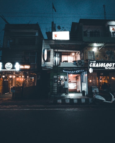 A nighttime street scene featuring a row of commercial buildings with illuminated signs. The buildings host various businesses, including a restaurant named Mr. IDLI and a tea shop named CHAiOLOGY. A person is seen entering one of the establishments, and there is a scooter parked on the street.