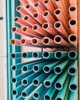 A group of multicolored pipes arranged vertically, with the top half in pink hues and the bottom half in teal. The pipes are tightly packed, creating a dynamic, industrial aesthetic.