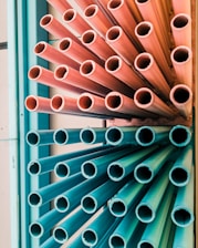 A group of multicolored pipes arranged vertically, with the top half in pink hues and the bottom half in teal. The pipes are tightly packed, creating a dynamic, industrial aesthetic.