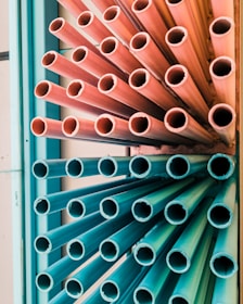 A group of multicolored pipes arranged vertically, with the top half in pink hues and the bottom half in teal. The pipes are tightly packed, creating a dynamic, industrial aesthetic.