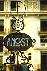 A rusted and ornate iron sign with the word 'ANGST' is centered in front of an old, decaying building with broken windows. The building's facade shows signs of wear, with faded paint and structural deterioration.