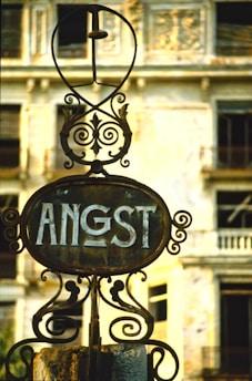 A rusted and ornate iron sign with the word 'ANGST' is centered in front of an old, decaying building with broken windows. The building's facade shows signs of wear, with faded paint and structural deterioration.