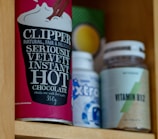 A cupboard shelf holds a variety of packaged goods. On the left, there is a red container of Clipper hot chocolate with bold, white lettering. Next to it, there’s a blue and white bottle, partially obscured, and a vitamin B12 supplement bottle with a green and beige label.