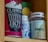 A cupboard shelf holds a variety of packaged goods. On the left, there is a red container of Clipper hot chocolate with bold, white lettering. Next to it, there’s a blue and white bottle, partially obscured, and a vitamin B12 supplement bottle with a green and beige label.