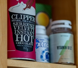 A cupboard shelf holds a variety of packaged goods. On the left, there is a red container of Clipper hot chocolate with bold, white lettering. Next to it, there’s a blue and white bottle, partially obscured, and a vitamin B12 supplement bottle with a green and beige label.