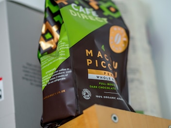 A package of whole bean coffee from Café Direct, featuring a green and brown design with text indicating that it is organic and from Peru, specifically named Machu Picchu. The packaging also includes details on its flavor profile, such as full-bodied and dark chocolate.