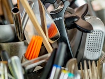 A collection of kitchen utensils and tools is densely packed, including a grater, knife sharpener, wooden spoon, and silicone brush. The utensils are made of metal, plastic, and wood, with some items overlapping each other.