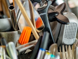 A collection of kitchen utensils and tools is densely packed, including a grater, knife sharpener, wooden spoon, and silicone brush. The utensils are made of metal, plastic, and wood, with some items overlapping each other.