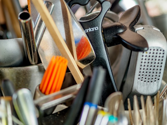 A collection of kitchen utensils and tools is densely packed, including a grater, knife sharpener, wooden spoon, and silicone brush. The utensils are made of metal, plastic, and wood, with some items overlapping each other.