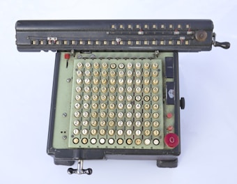 An vintage mechanical calculator featuring a large number pad with keys labeled with numbers ranging from 0 to 9. The device has a hand crank on the side and a series of switches and levers on the top panel. The body is predominantly black with a mint green section where the keys are located. Some red and black buttons are also visible, contributing to its complex appearance.